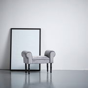 Grauer Hocker mit gepolsterten Armlehnen und schwarzen Beinen, seitliche Perspektive, vor einem großen Spiegel aufgestellt.