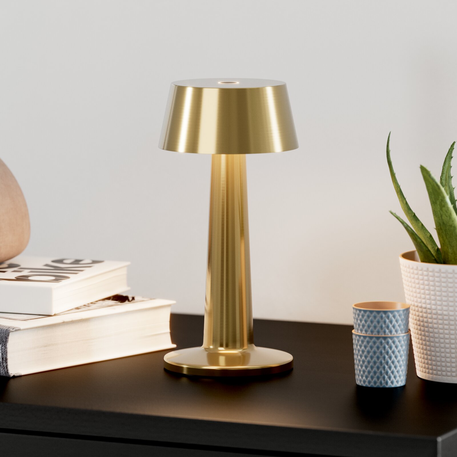 Goudkleurige metalen tafellamp met conische kap en ronde voet op een donkere tafel, vooraanzicht op ooghoogte ernaast een stapel boeken, twee blauwe bekers en een potplant tegen een lichte achtergrond