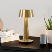 Goudkleurige metalen tafellamp met conische kap en ronde voet op een donkere tafel, vooraanzicht op ooghoogte; ernaast een stapel boeken, twee blauwe bekers en een potplant tegen een lichte achtergrond