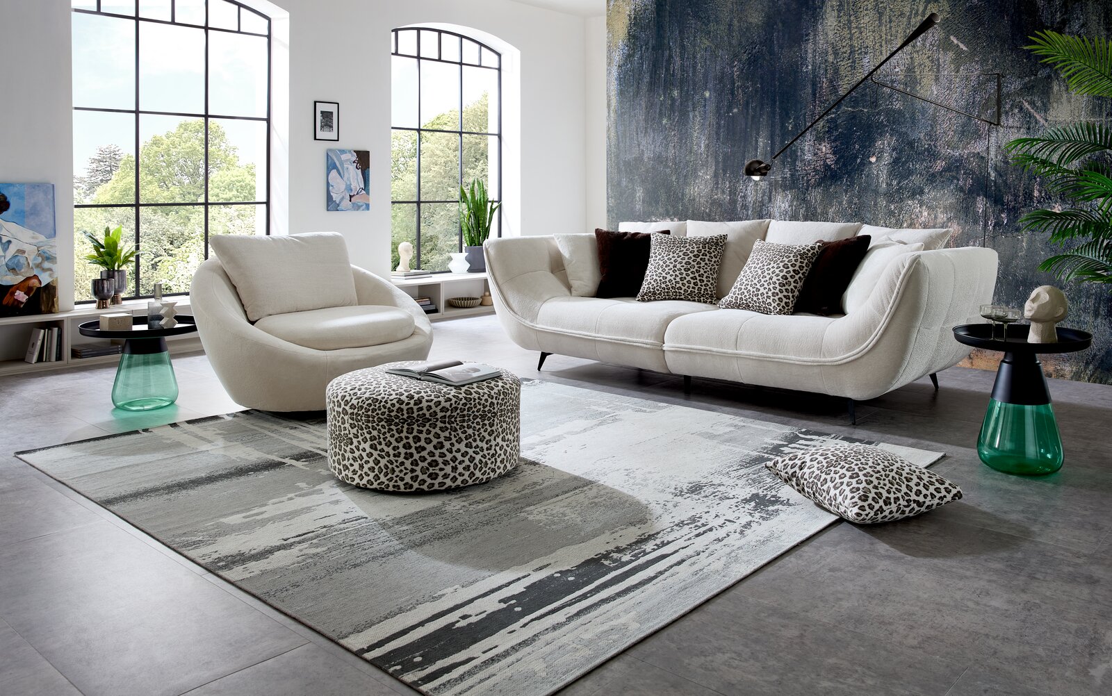 Modernes Wohnzimmer-Set mit cremefarbenem Sofa und passendem Sessel, Leopardmuster-Pouf und Kissen auf großem grauen Teppich vor großer Fensterfront; Perspektive: frontale Weitwinkelaufnahme auf Augenhöhe.