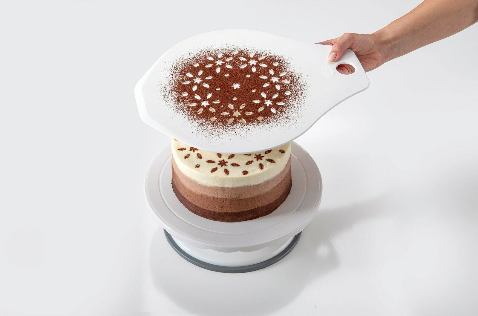 Hand hält einen Tortenretter mit Schablone über einer mehrschichtigen Torte, um ein dekoratives Muster mit Kakaopulver zu erzeugen. Perspektive von oben.