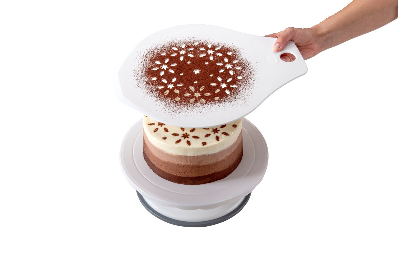 Hand hält einen Tortenretter mit Schablone über einer mehrschichtigen Torte, um ein dekoratives Muster mit Kakaopulver zu erzeugen. Perspektive von oben.