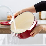 Roter runder Gusseisen-Bräter von KitchenAid wird in einem Waschbecken unter fließendem Wasser gereinigt, seitliche Perspektive.