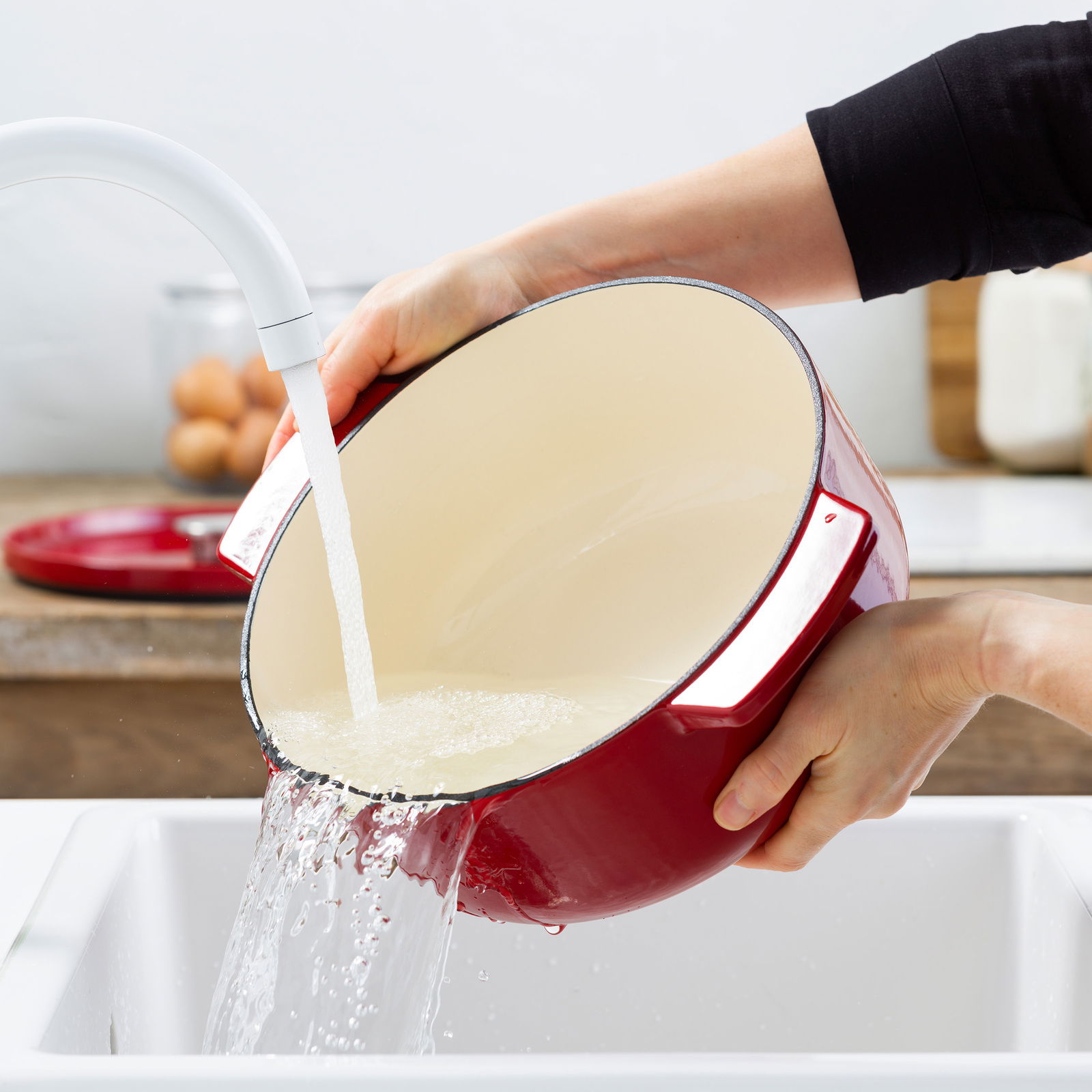 Roter runder Gusseisen-Bräter von KitchenAid wird in einem Waschbecken unter fließendem Wasser gereinigt, seitliche Perspektive.