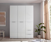 Weißer, moderner Kleiderschrank mit drei Türen und zwei Schubladen, frontal in einem hellen Schlafzimmer fotografiert; rechts Fenster mit Vorhang und Zimmerpflanze.