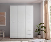 Weißer, moderner Kleiderschrank mit drei Türen und zwei Schubladen, frontal in einem hellen Schlafzimmer fotografiert; rechts Fenster mit Vorhang und Zimmerpflanze.
