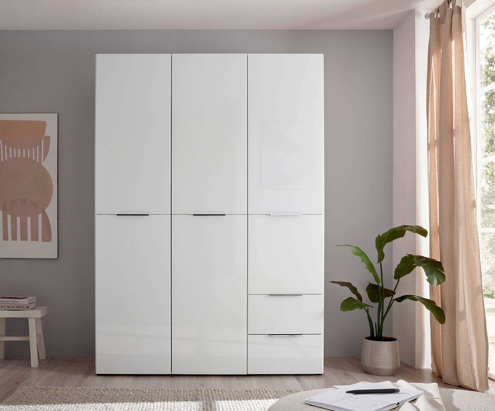 Weißer, moderner Kleiderschrank mit drei Türen und zwei Schubladen, frontal in einem hellen Schlafzimmer fotografiert; rechts Fenster mit Vorhang und Zimmerpflanze.