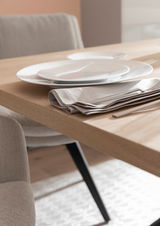 Nahaufnahme eines gedeckten Esstisches mit Holzoberfläche, weißen Tellern und beigen Stoffservietten, seitliche Perspektive