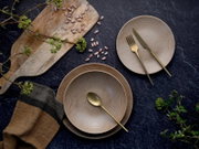 Draufsicht auf ein rustikales Tisch-Arrangement mit zwei beigefarbenen Keramiktellern, goldenem Besteck, Holzbrett, gestreuten Bohnen und grünen Zweigen auf dunkler Steinoberfläche