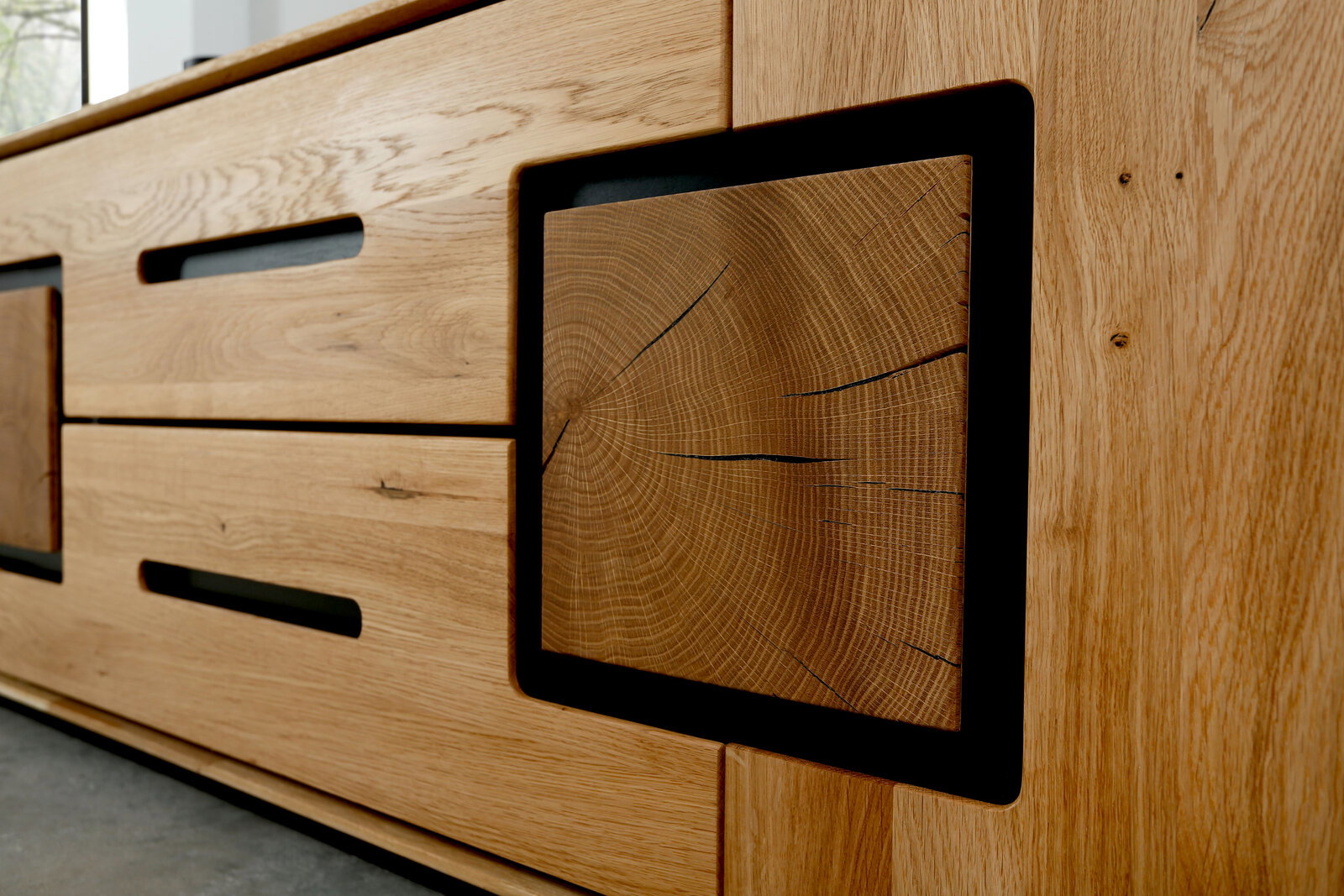 Detailaufnahme eines modernen Sideboards aus hellem Eichenholz mit sichtbarer Maserung, schwarzen Griffaussparungen und dekorativem Stirnholz-Element im schwarzen Rahmen, fotografiert aus schräger Frontperspektive