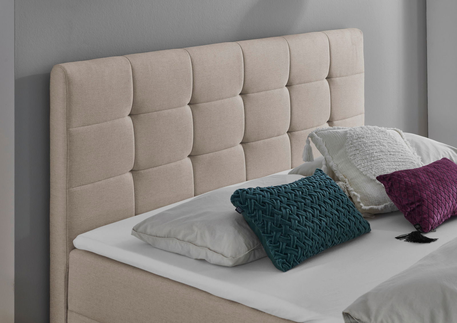 Beige gepolstertes Boxbett mit gestepptem Kopfteil, dekoriert mit Kissen in verschiedenen Farben, seitliche Perspektive.
