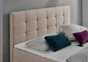 Beige gepolstertes Boxbett mit gestepptem Kopfteil, dekoriert mit Kissen in verschiedenen Farben, seitliche Perspektive.