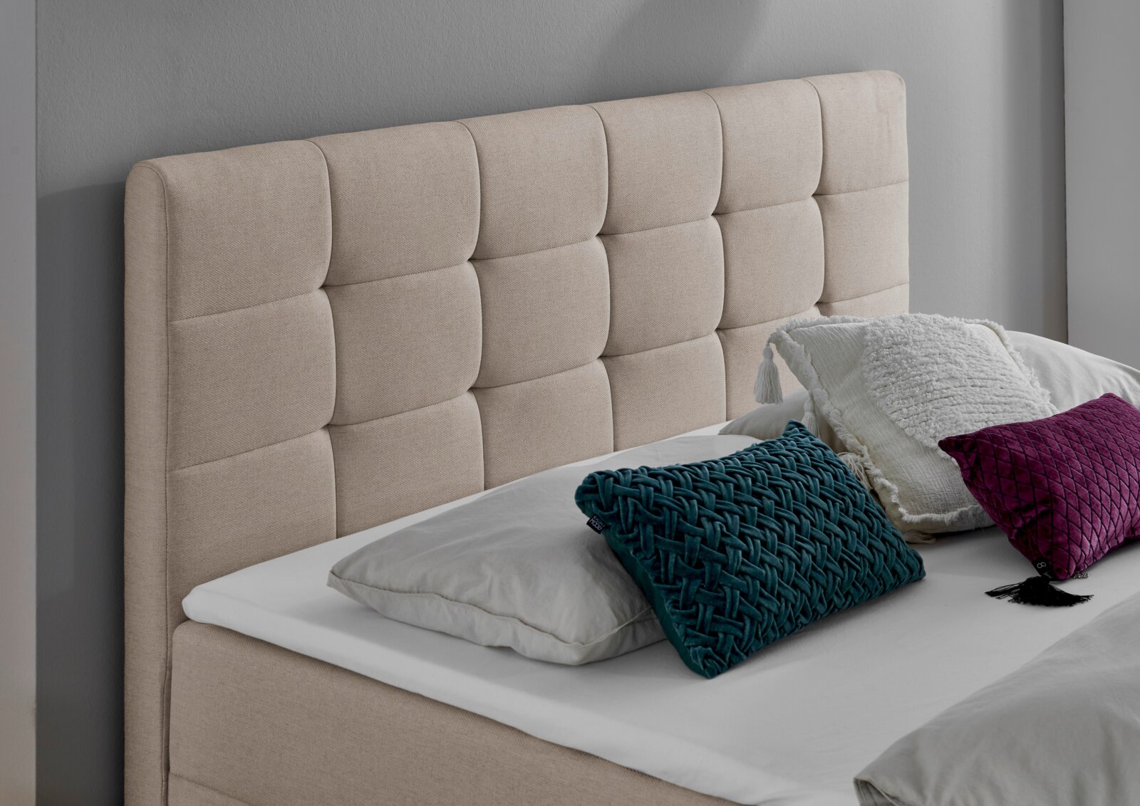 Beige gestoffeerd boxbed met doorgestikt hoofdeinde, versierd met kussens in verschillende kleuren, zijaanzicht.