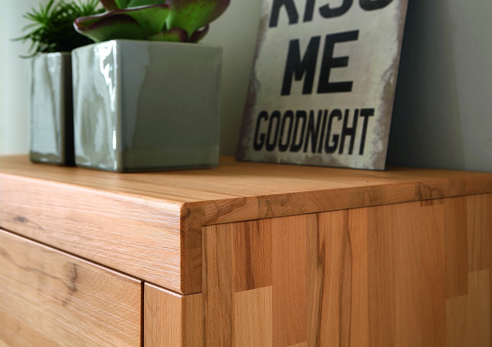 Nahaufnahme eines Highboards aus Holz mit dekorativen Elementen wie Pflanzen in Keramiktöpfen und einem Schild mit der Aufschrift 'Kiss Me Goodnight'.