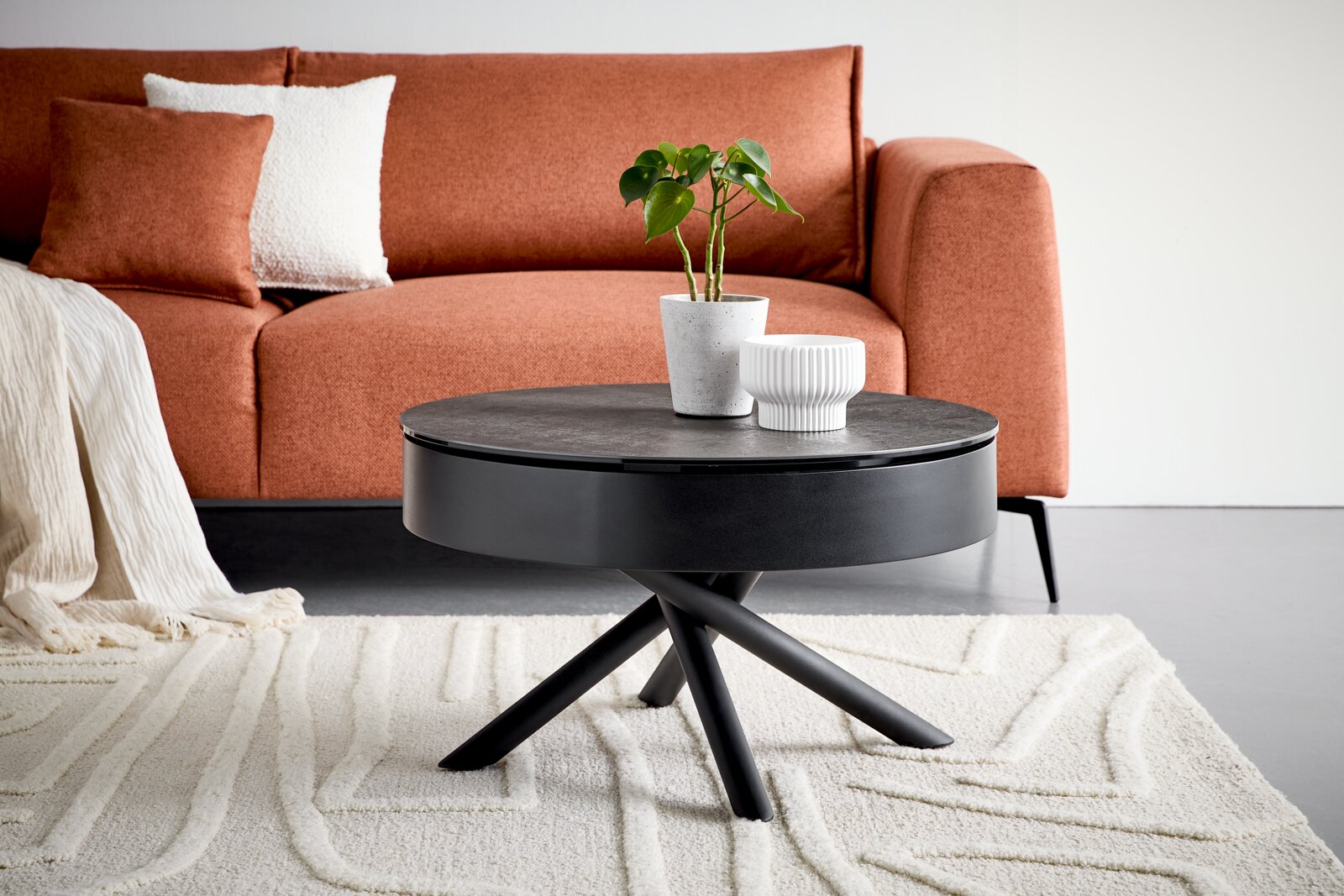 Novel Couchtisch STROMBOLI Runder schwarzer Couchtisch mit dunkler Tischplatte, breitem Korpus und gekreuztem Metallgestell, aus leichter Frontalperspektive in einem modernen Wohnzimmer vor einem terrakottafarbenen Sofa gezeigt