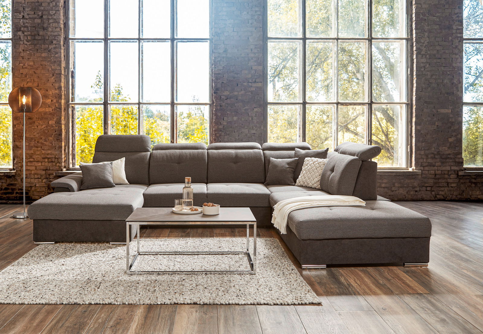 Großes, modernes graues Ecksofa in einem hellen Raum mit großen Fenstern, frontal betrachtet.