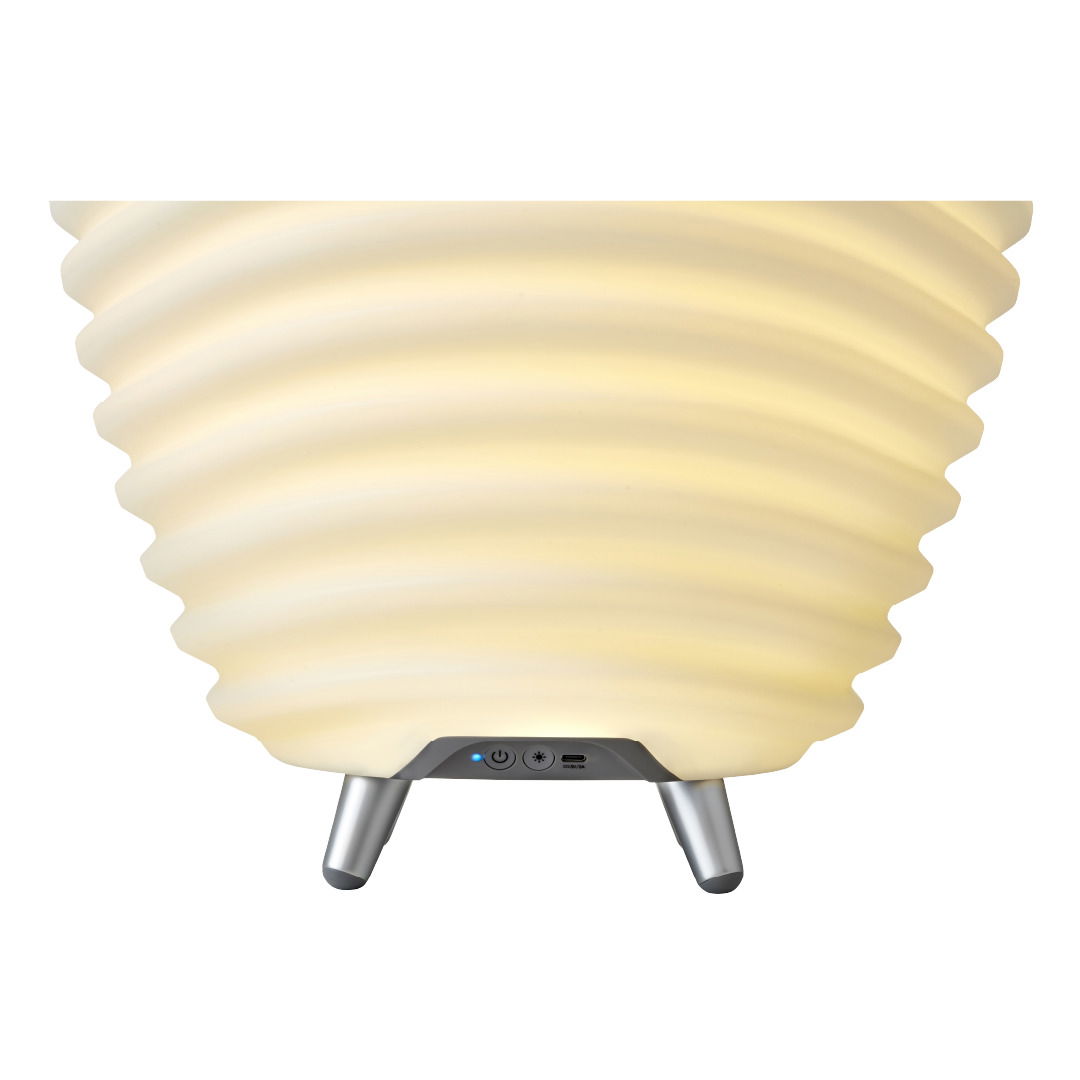 Nahaufnahme einer Akku-Leuchte mit Lautsprecher, die auf drei Beinen steht. Die Oberfläche ist geriffelt und leuchtet in einem warmen Gelbton.