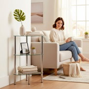 Dreistufiges Glas-Regal mit verchromten Metallbeinen neben einem hellen Sofa im Wohnzimmer, seitliche Frontansicht auf das Produkt.