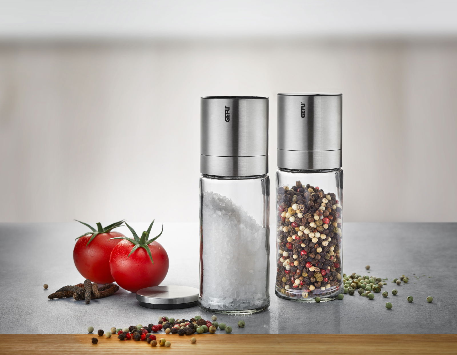 Zwei Salz- und Pfeffermühlen LAMOLA aus Glas und Edelstahl, gefüllt mit grobem Salz und buntem Pfeffer, auf einer Küchenarbeitsplatte. Perspektive: Frontalansicht.
