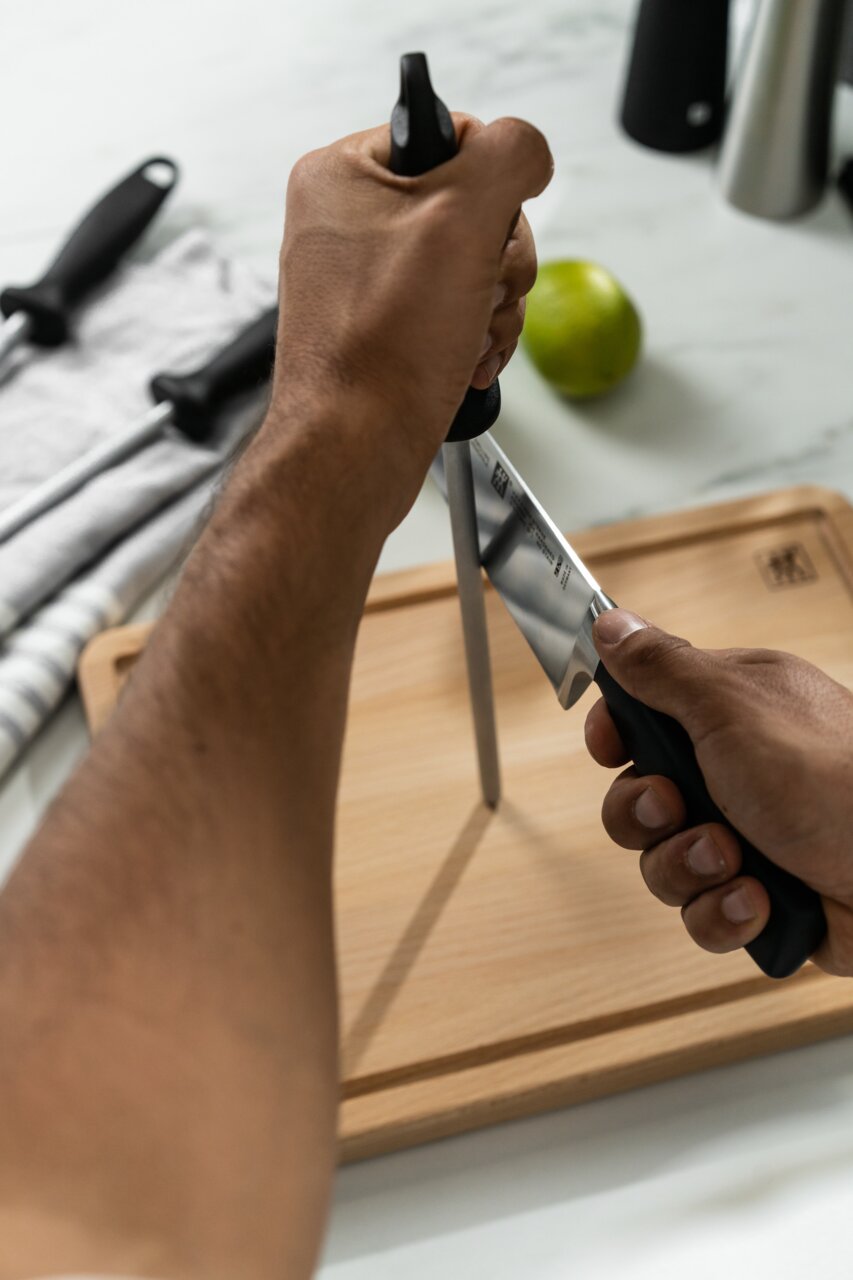 Person schärft ein Messer mit einem 23 cm langen Wetzstahl auf einem Holzbrett, seitliche Perspektive.