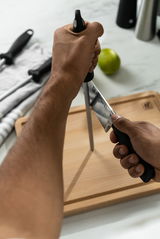 Person schärft ein Messer mit einem 23 cm langen Wetzstahl auf einem Holzbrett, seitliche Perspektive.