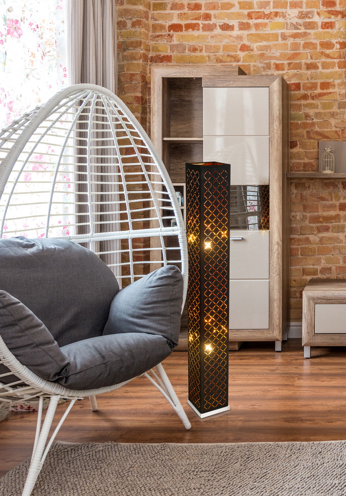 Moderne Stehleuchte aus Holz mit geometrischem Muster, beleuchtet in einem gemütlichen Wohnzimmer mit Ziegelwand und Hängesessel, seitliche Perspektive.