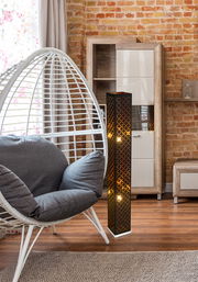 Moderne Stehleuchte aus Holz mit geometrischem Muster, beleuchtet in einem gemütlichen Wohnzimmer mit Ziegelwand und Hängesessel, seitliche Perspektive.