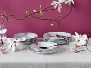 Schlichtes weißes Geschirrset mit rosa Blütenmotiv (Teller und Schalen) auf gedecktem Tisch, dekoriert mit Magnolienzweigen vor pinkem Hintergrund, fotografiert in frontaler leicht erhöhter Perspektive.