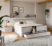 Weißes Holzbett im Landhausstil mit naturfarbenen Akzenten und ausgezogener Bettkasten-Schublade, in der ein Hund sitzt; Perspektive: frontale Ansicht leicht von links in einem hellen Schlafzimmer.