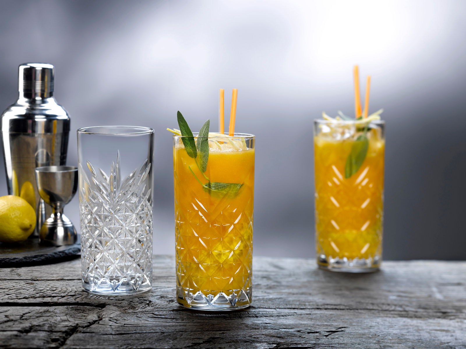 Drei Gläser aus dem Glasset Timeless 8-tlg. auf einem Holztisch, zwei gefüllt mit einem gelben Getränk und Dekoration, im Hintergrund ein Cocktailshaker und Zitronen, seitliche Perspektive.