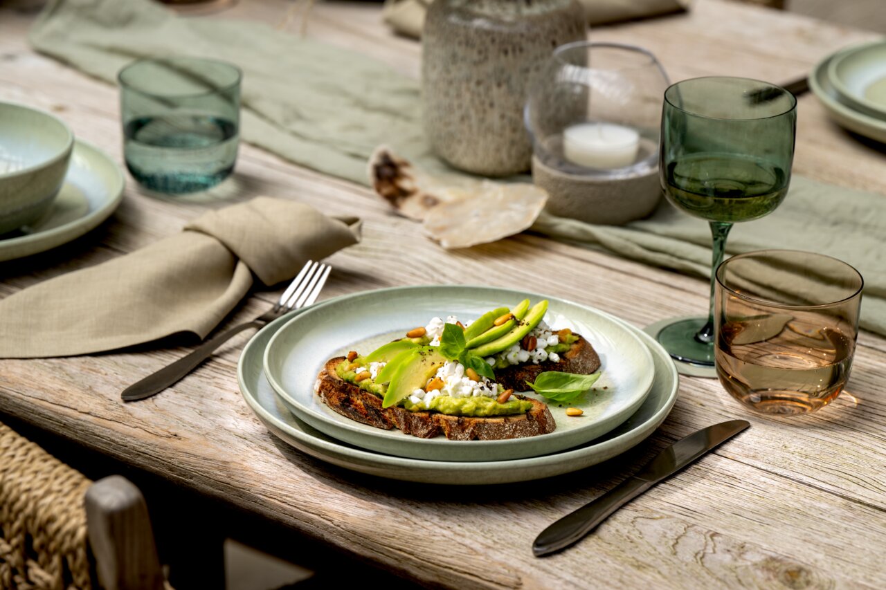 Ein gedeckter Tisch mit einem grünen Speiseteller, darauf Avocado-Toast mit Hüttenkäse und Pinienkernen. Perspektive von schräg oben.