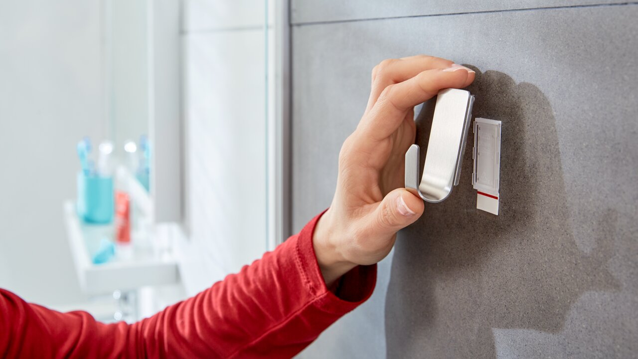 Hand befestigt einen tesa Powerstrip Waterproof Haken an einer Badezimmerwand, seitliche Perspektive.