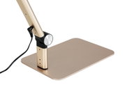 Nahaufnahme des Sockels einer goldfarbenen Schreibtischlampe mit rechteckiger Standplatte, Drehgelenk und angeschlossenem Stromkabel, schräg von der Seite aufgenommen.
