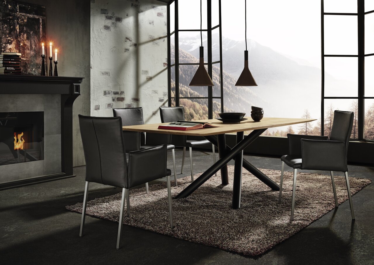 WK-Wohnen Esstisch (240) WK 852 Moderner Esstisch mit Holzplatte und schwarzen Metallbeinen, umgeben von vier schwarzen Stühlen, in einem stilvollen Raum mit Kamin und Panoramafenster. Perspektive von der Seite.