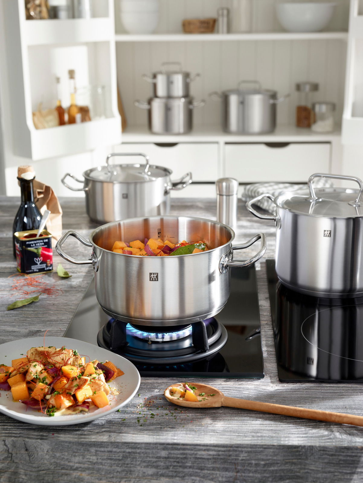 Küchenansicht mit einem Topfset aus Edelstahl, das auf einem Herd steht. Der vordere Topf ist mit Gemüse gefüllt und wird von der Seite gezeigt.