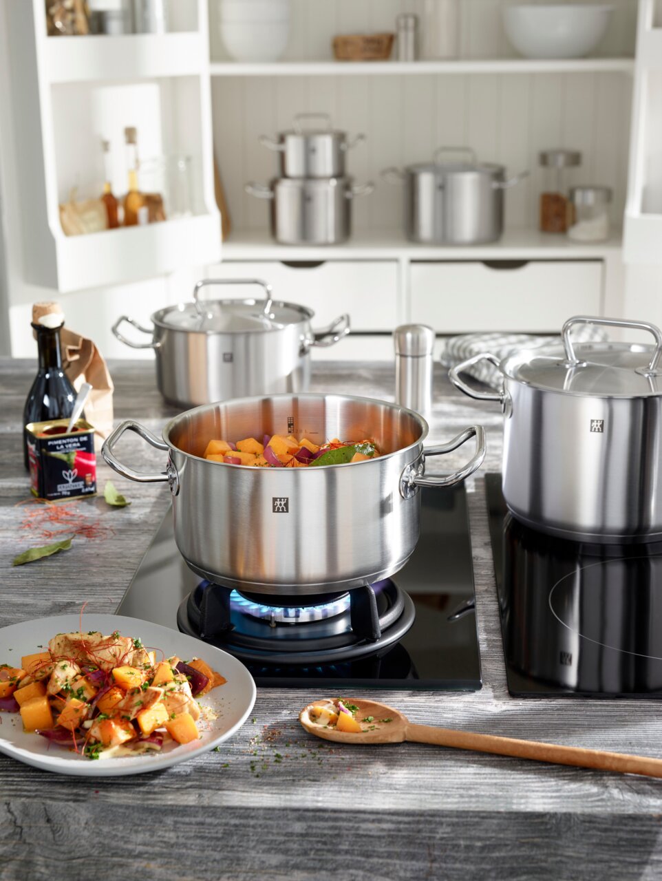 Küchenansicht mit einem Topfset aus Edelstahl, das auf einem Herd steht. Der vordere Topf ist mit Gemüse gefüllt und wird von der Seite gezeigt.