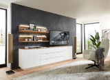 Modernes Wohnzimmer mit Wandboard CORE, bestehend aus zwei hölzernen Regalböden an einer dunklen Wand, dekoriert mit Pflanzen und Büchern, im Hintergrund ein großer Fernseher auf einem weißen Sideboard, seitliche Perspektive.