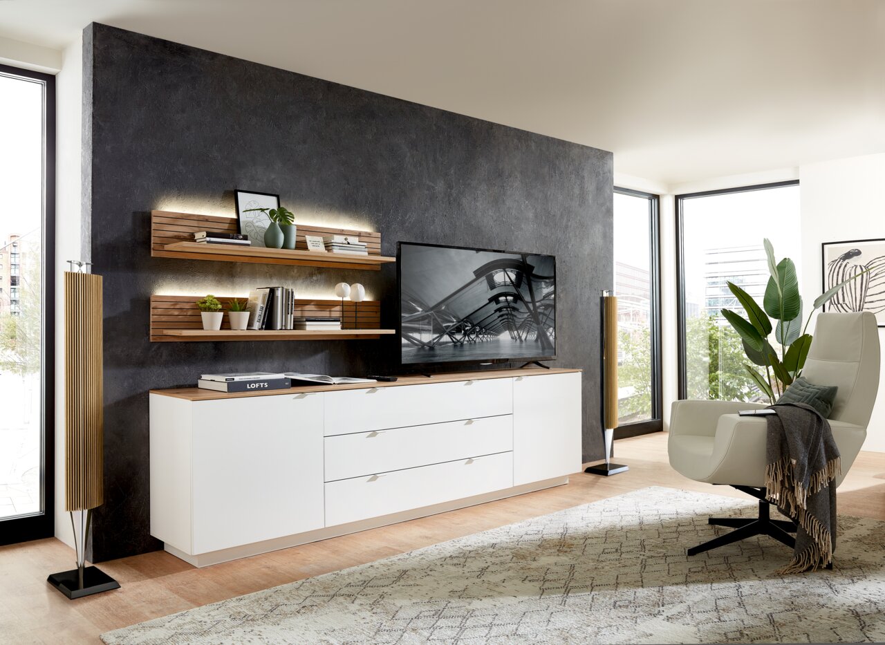 Modernes Wohnzimmer mit Wandboard CORE, bestehend aus zwei hölzernen Regalböden an einer dunklen Wand, dekoriert mit Pflanzen und Büchern, im Hintergrund ein großer Fernseher auf einem weißen Sideboard, seitliche Perspektive.