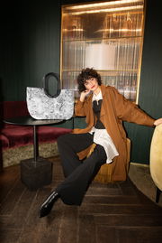 Stilvolle Frau in einem eleganten Raum, sitzend neben einer großen, gemusterten Tasche mit runden Griffen auf einem Tisch. Die Tasche ist im Vordergrund sichtbar.