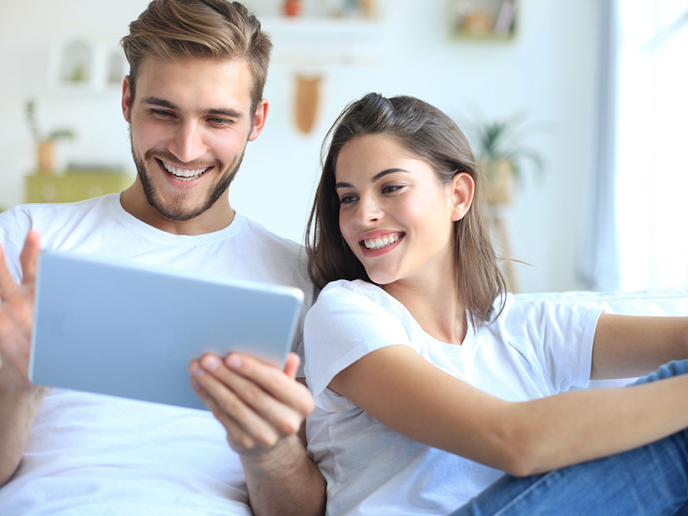 Ein Paar in weißen Shirts sitzt auf dem Sofa und schaut lächelnd zusammen auf ein Tablet.