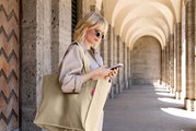 Beige gerippte Stoff-Shopper-Tasche mit langen Henkeln und rotem Label, getragen über der Schulter einer Frau in einer Arkadenpassage, seitliche Perspektive