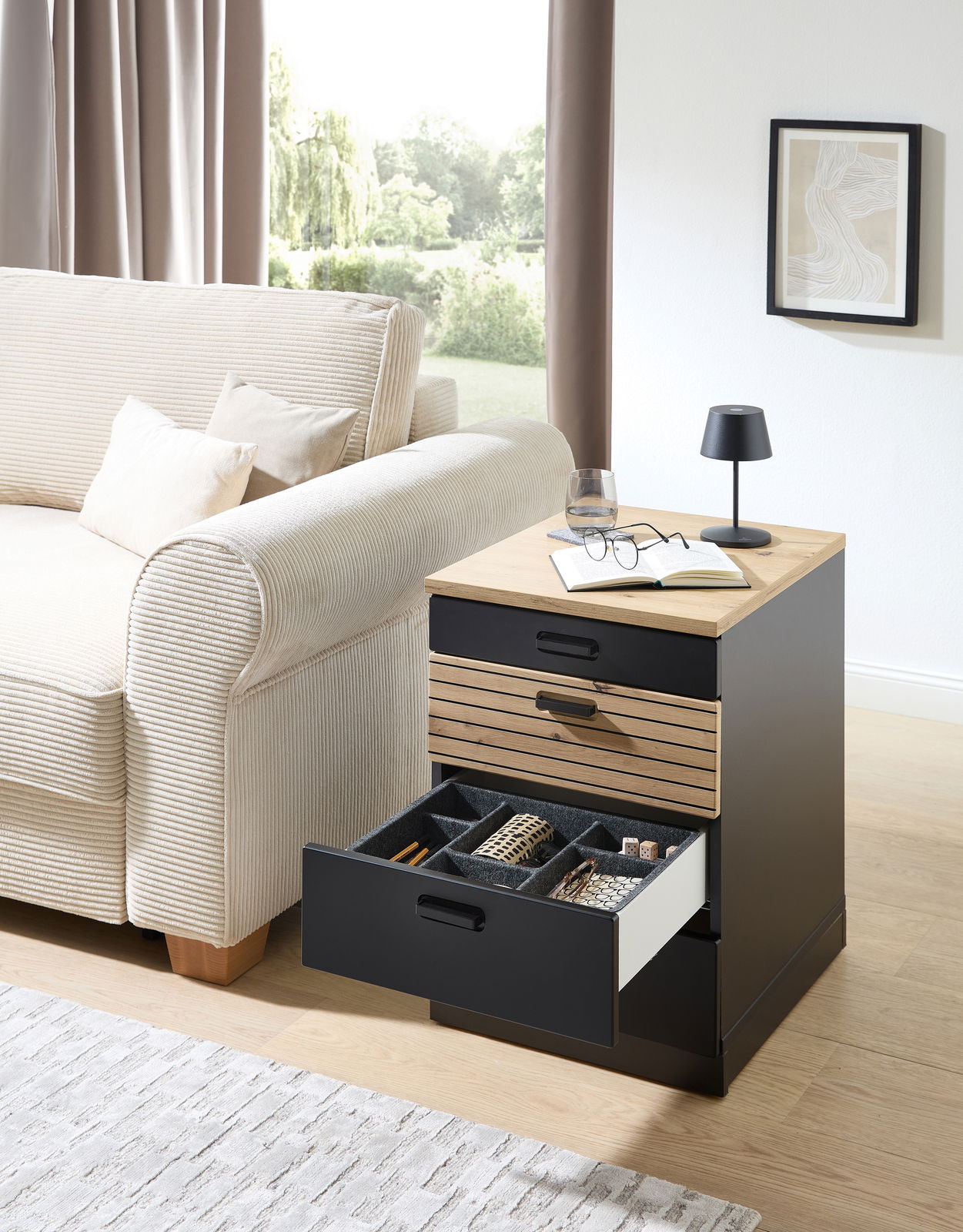 Rollcontainer mit offenem Schubfach, gefüllt mit Accessoires, neben einem Sofa in einem Wohnzimmer, aus seitlicher Perspektive fotografiert.