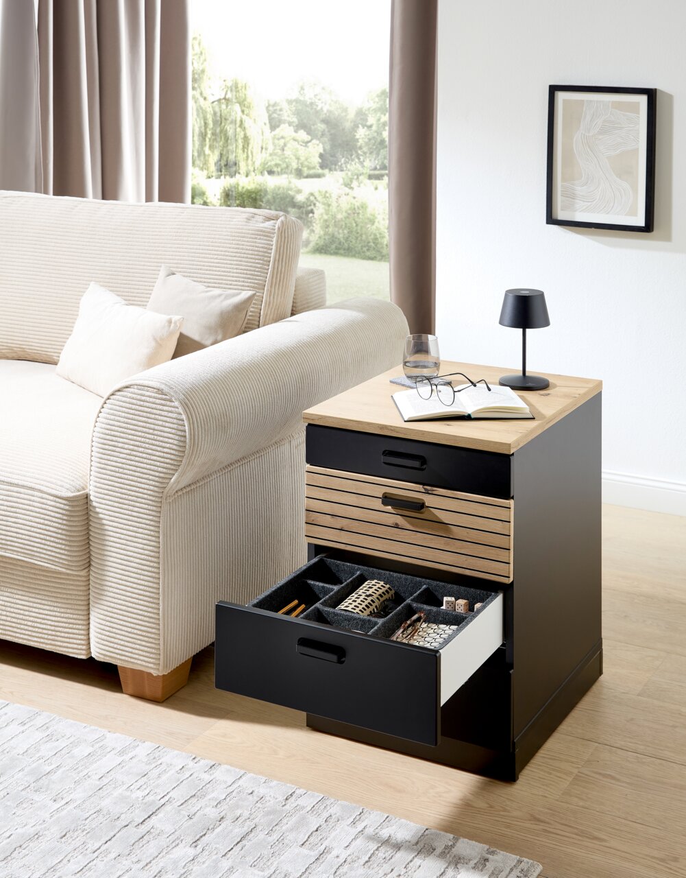 Rollcontainer mit offenem Schubfach, gefüllt mit Accessoires, neben einem Sofa in einem Wohnzimmer, aus seitlicher Perspektive fotografiert.