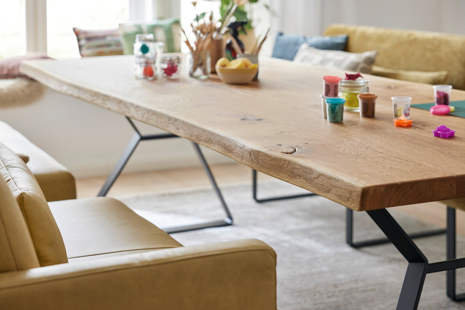 Holzesstisch mit natürlicher Kante und Metallbeinen, seitliche Perspektive, dekoriert mit Bastelmaterialien und umgeben von beigen Sofas.