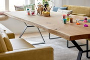 Holzesstisch mit natürlicher Kante und Metallbeinen, seitliche Perspektive, dekoriert mit Bastelmaterialien und umgeben von beigen Sofas.
