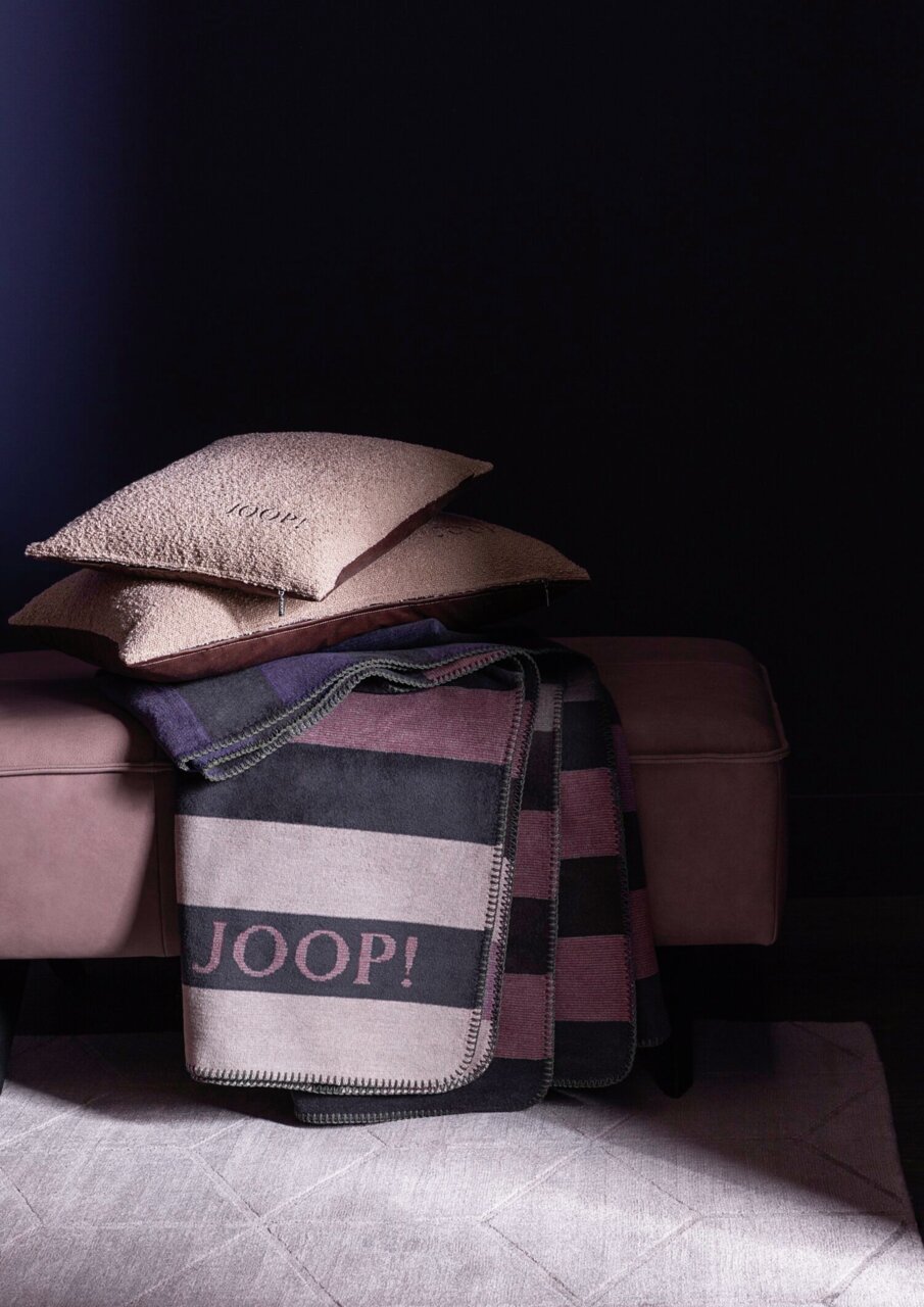 Zwei beige Zierkissen mit der Aufschrift 'JOOP!' auf einem Sofa, daneben eine gestreifte Decke mit dem gleichen Schriftzug. Die Perspektive ist seitlich.