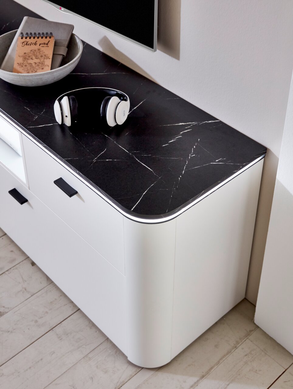 Close-up van een modern wit dressoir met afgeronde hoeken en een zwart marmeren blad, met daarop een koptelefoon en een notitieboekje.