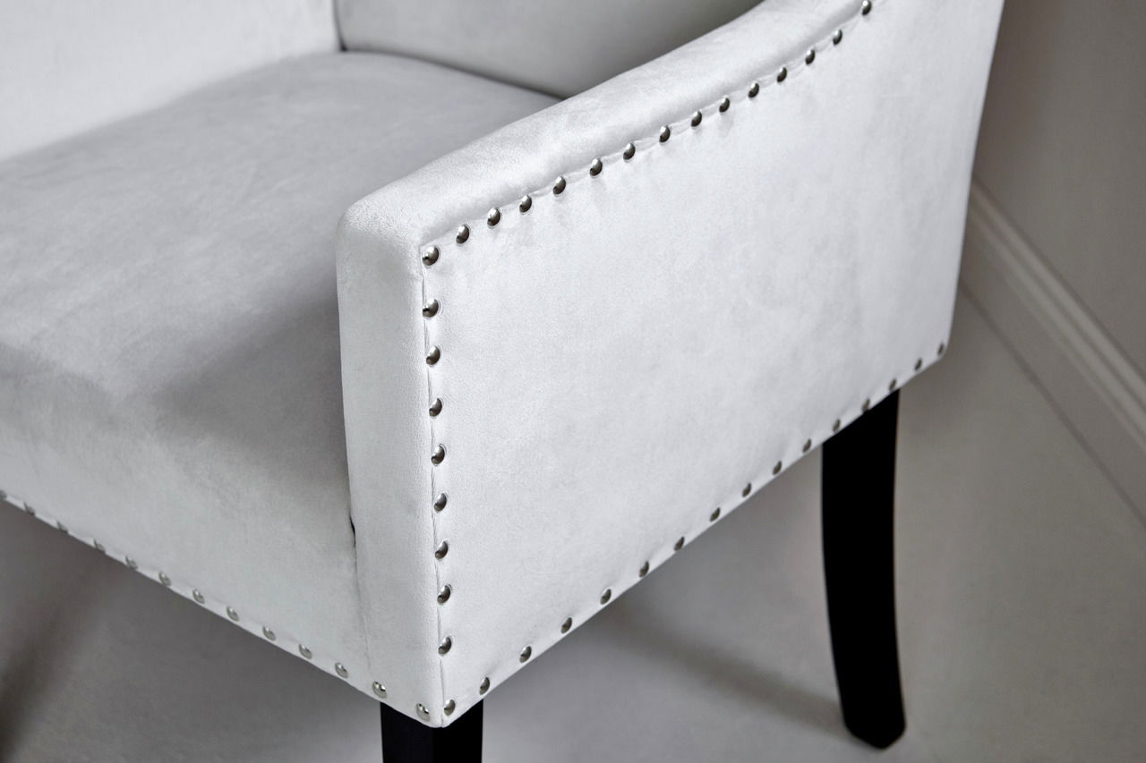 Nahaufnahme eines eleganten, hellgrauen Stuhls mit Nietenverzierung und schwarzen Beinen, seitliche Perspektive.