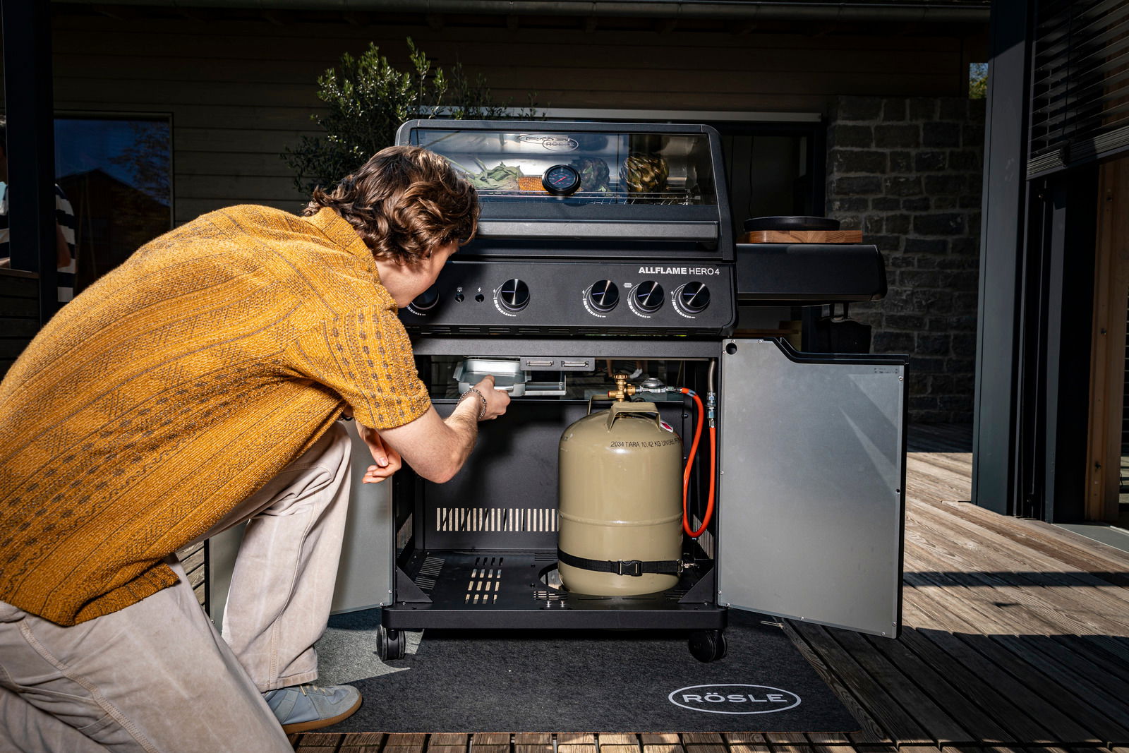 Mann überprüft Gasgrill Allflame Hero 4 von vorne, mit geöffneter Tür und sichtbarer Gasflasche.