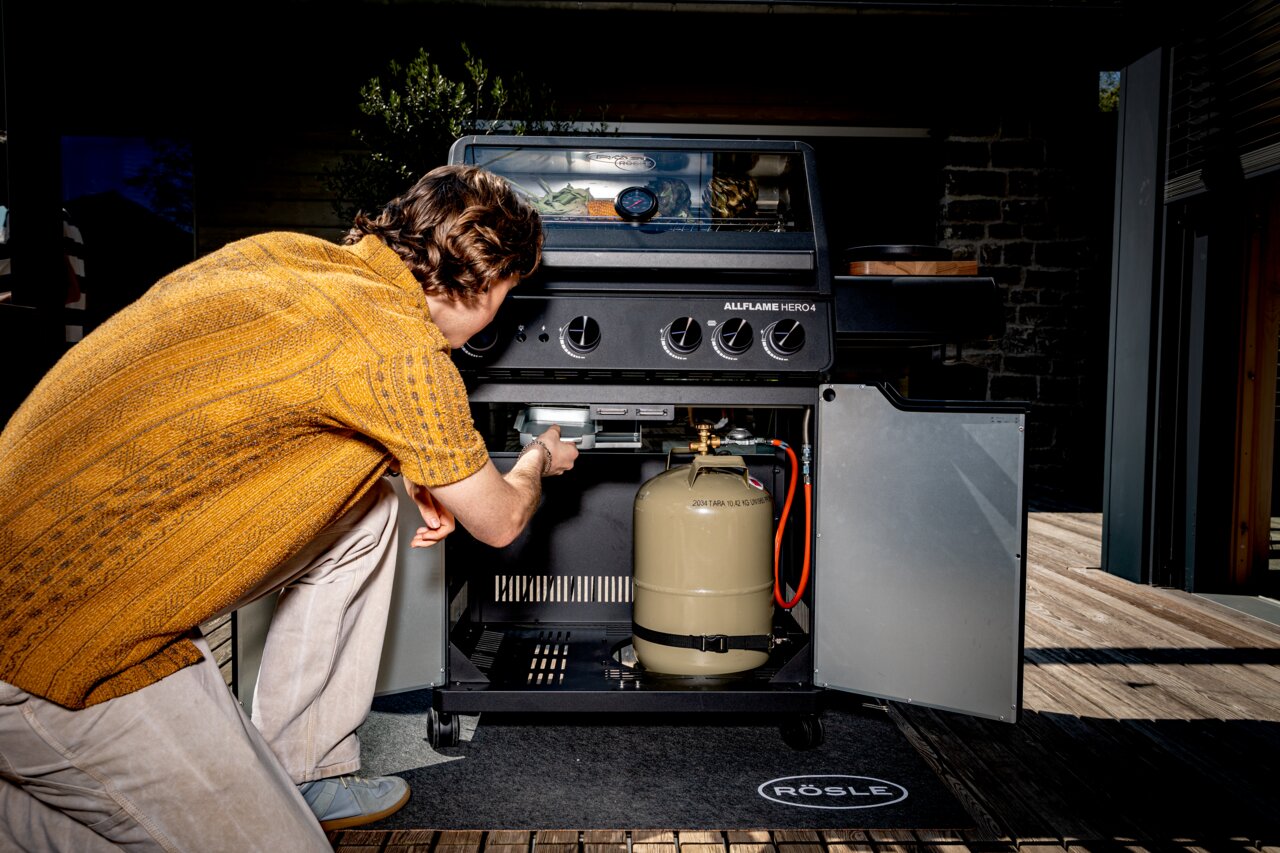 Een man bekijkt de Allflame Hero 4-gasbarbecue van voren, met geopende deur en zichtbare gasfles.